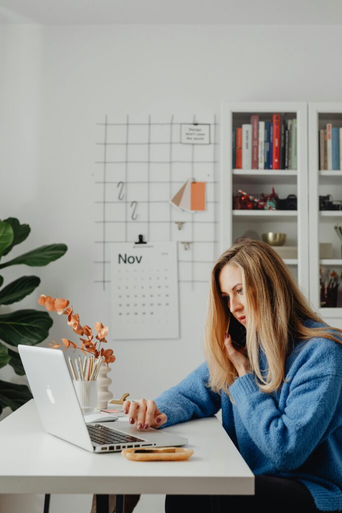 Woman multitasking at home office desk, using laptop and phone, surrounded by books and calendar.
