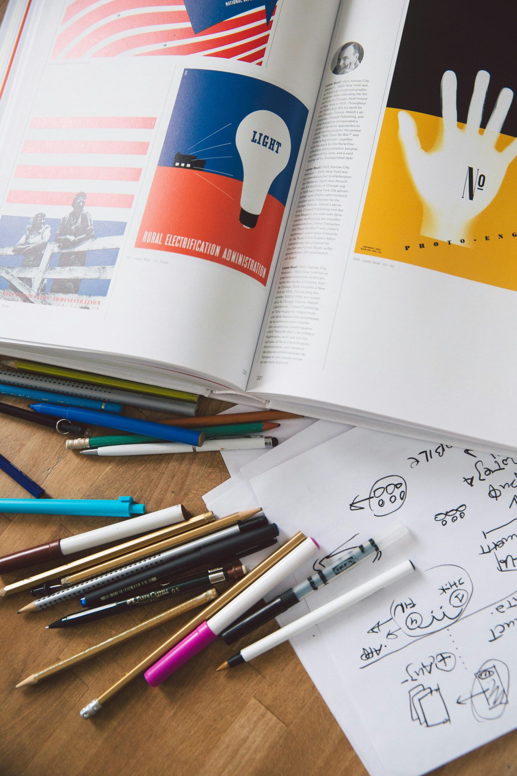 An artistic workspace featuring an open book, scattered pens, and handwritten sketches on a desk.